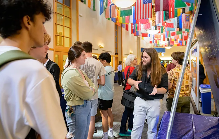 Student explains project to a small group of peers in a hallway decorated with international flags.