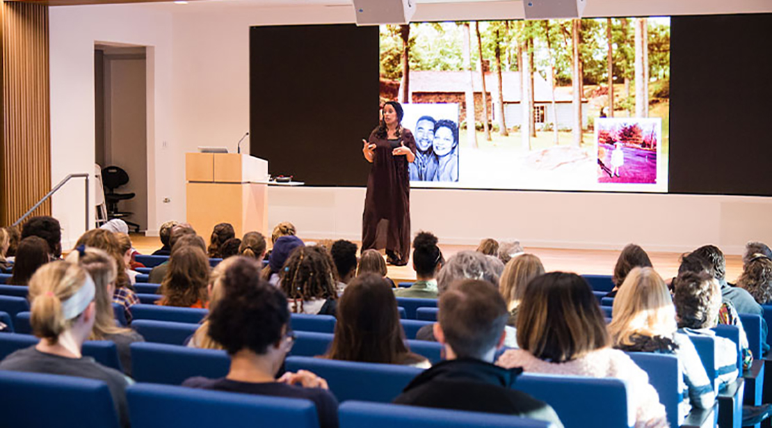 Woman wearing black gestures to audience while standing in front of a screen showing a house and picture of a couple; the auditorium is full of seated students.