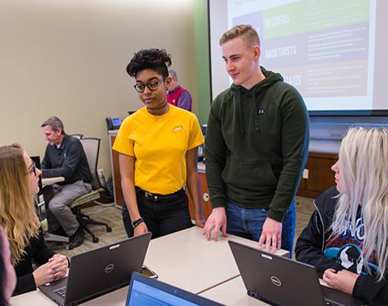 Two students stand at table of students with laptops