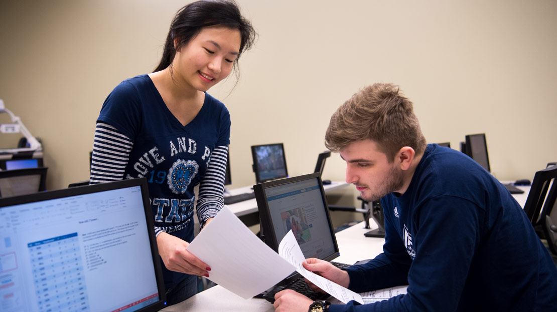 Two students sharing a piece of paper and working on a computer.