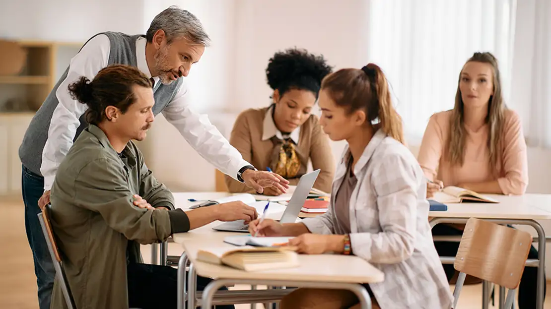 A class of adult students with professor assisting one of them.
