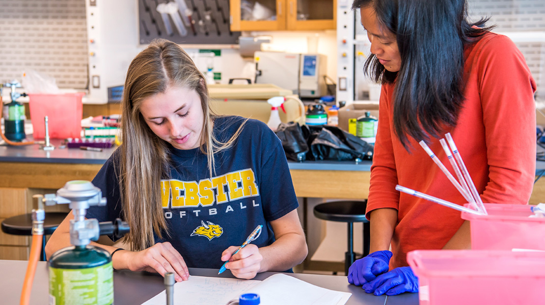 Student seated at lab table with professor leaning and talking with her.
