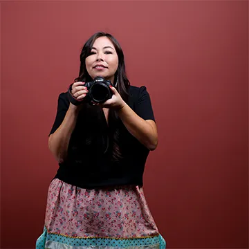 Matika Wilbur, self-portrait, stands against a red-toned background holding a camera up near her face.
