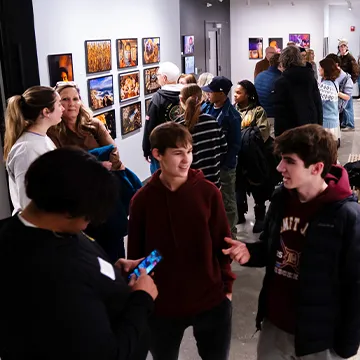 Exhibition hall full of student presenters, exhibit visitors and photography displayed on walls.