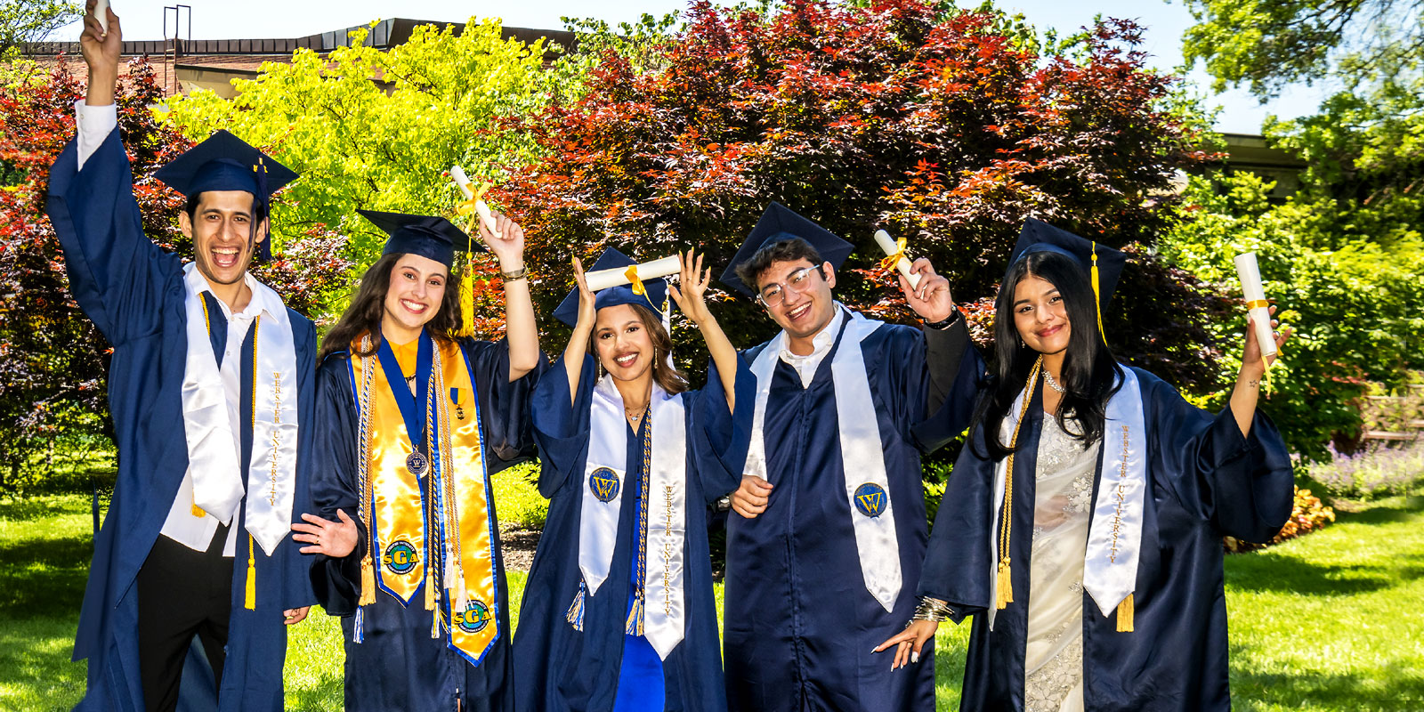 Group of celebrating graduates in cap and gown hold diplomas in air.