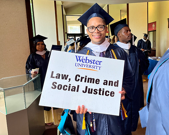A Webster student wearing commencement regalia holding a sign for their department that reads "Law, Crime and Social Justice".