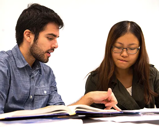 Two students work together at a table with workbooks and other school supplies