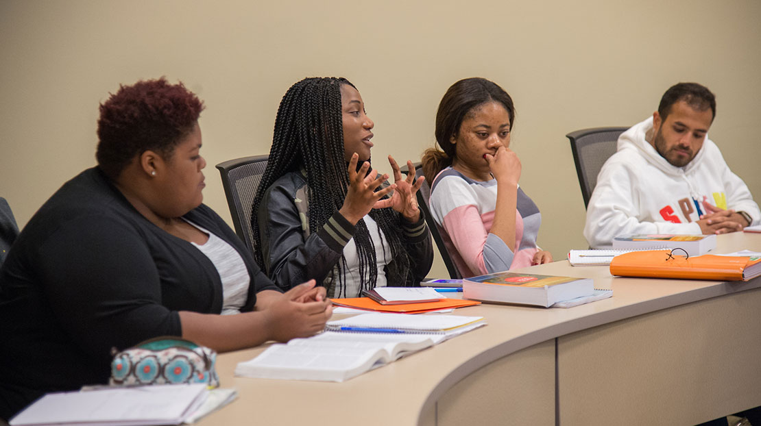 students at circular desk; one of them speaking to the group.