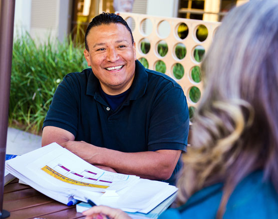 Smiling man with medium-skin-tone who is sitting at table with open notes in front of him talking to another person