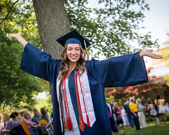 A student wearing a commencement robe with hands outstretched while smiling.