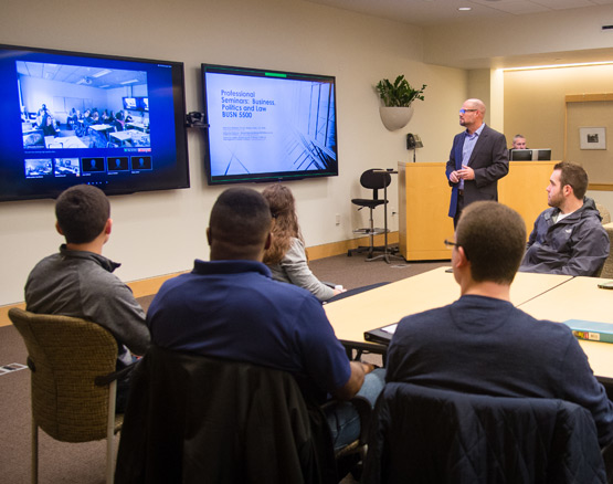 Professor and students in a classroom look at large screens with another classroom and course material